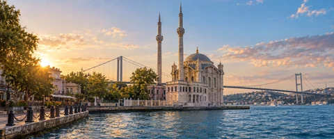 Destination artwork of Istanbul: Istanbul's Ortakoy Mosque by the Bosphorus, the suspension bridge behind, blue water and warm historic architecture in a geographically believable shoreline view.